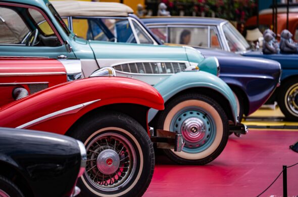 Four classic cars in black, red, teal, and blue are displayed at a car show. Photo by Peter Xie from Pexels
