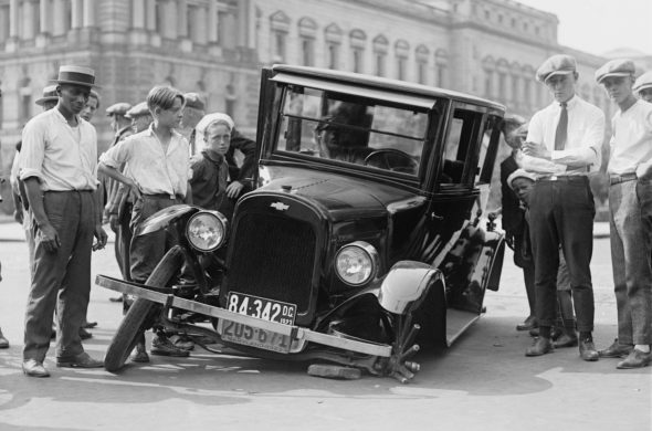 A black and white old photo of people posing beside a flat tired black car in the road