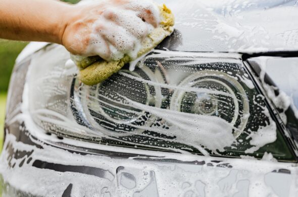 A hand holding a sponge washes the soapy headlights of a car Photo by KaboomPics from Pexel