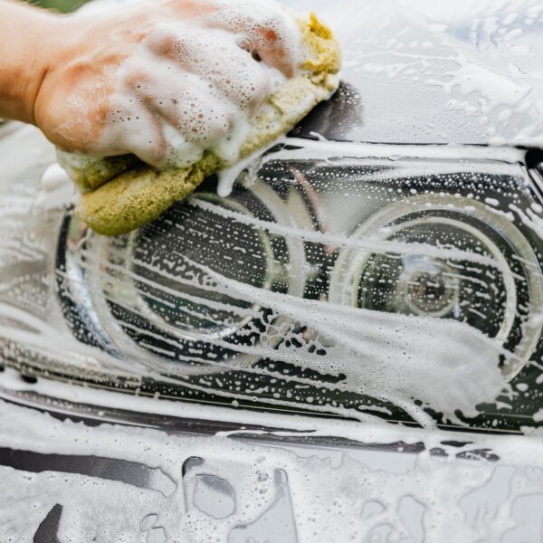 A hand holding a sponge washes the soapy headlights of a car Photo by KaboomPics from Pexel