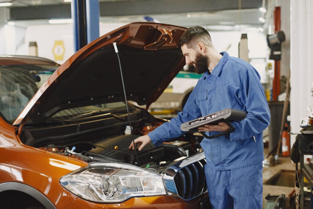 A mechanic inspects under the hood of an orange car. Photo by Gustavo Fring from Pexels