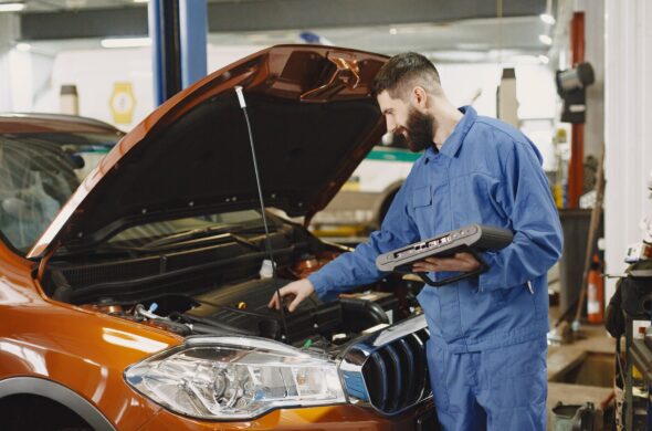 A mechanic inspects under the hood of an orange car. Photo by Gustavo Fring from Pexels