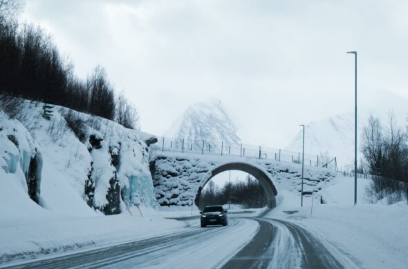 A car drives in winter on recently plowed streets under a bridge covered in snow. In the distance, snowy mountains are covered by fog. Photo by Utkarsh Malviya from Pexels