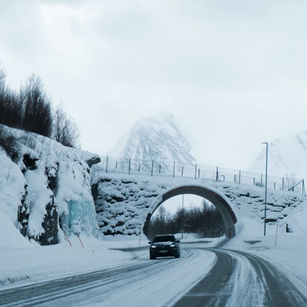 A car drives in winter on recently plowed streets under a bridge covered in snow. In the distance, snowy mountains are covered by fog. Photo by Utkarsh Malviya from Pexels