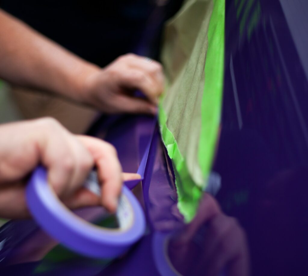 Two hands tape up a purple car with green and purple tape as they prepare for painting. Photo by Clarity Coat from Unsplash