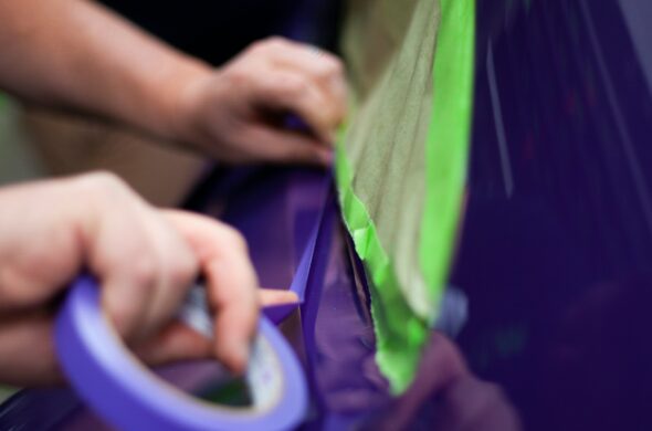 Two hands tape up a purple car with green and purple tape as they prepare for painting. Photo by Clarity Coat from Unsplash
