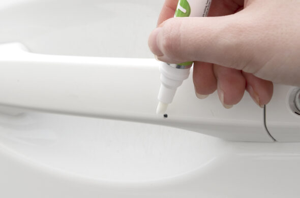 A hand touches up a chip on a white car door handle with a TouchUpDirect pen