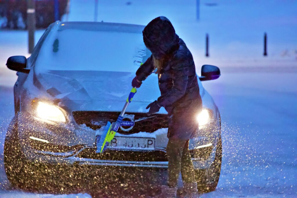A heavily bundled up woman in a snow storm brushes away snow from the front of her car with a brush. Photo by Gaith Shalan from Unsplash