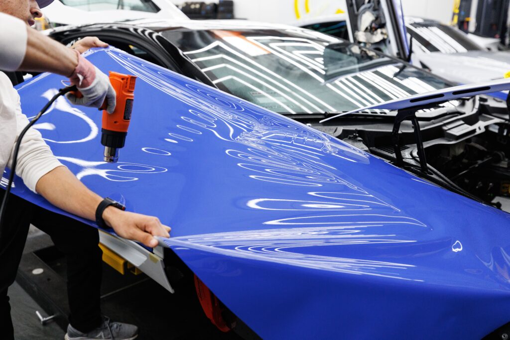 A technician uses a heat gun to carefully apply a blue vinyl wrap to a car in an auto workshop. Photo by Auto Records from Pexels