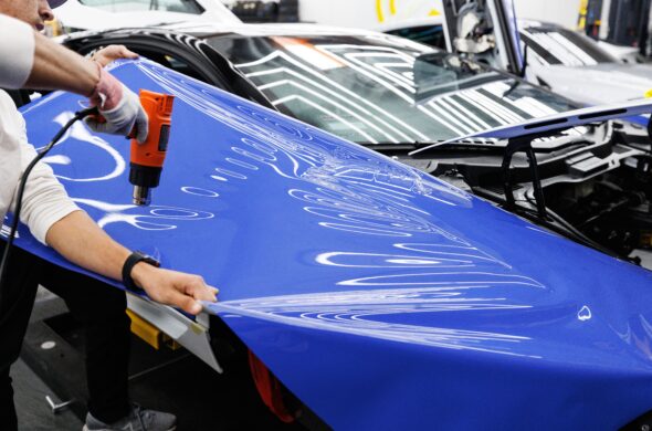 A technician uses a heat gun to carefully apply a blue vinyl wrap to a car in an auto workshop. Photo by Auto Records from Pexels
