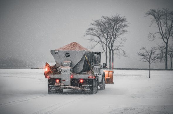 A snowplow truck spreading salt on a snowy road during a winter storm, with bare trees in the background. Photo by Cara Denison from Pexels