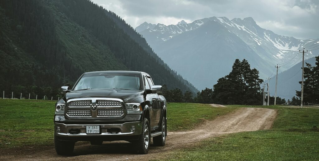 A Black Dodge Ram 1500 parked on a dirt road between green fields, with snow-capped mountains looming under a cloudy sky. Photo by Sergey Chuprin from Unsplash