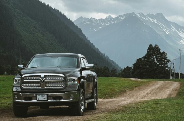 A Black Dodge Ram 1500 parked on a dirt road between green fields, with snow-capped mountains looming under a cloudy sky. Photo by Sergey Chuprin from Unsplash
