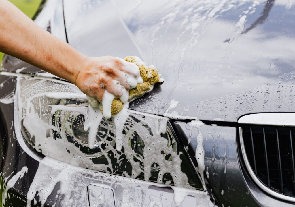 A hand washes the front of a black car using soap, water, and a sponge.
Photo by Kaboompics from Pexels