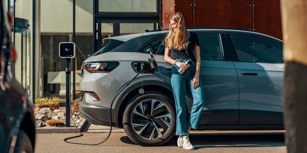 A girl leans against her silver EV as it charges at a station
Photo by JUICE from Unsplash